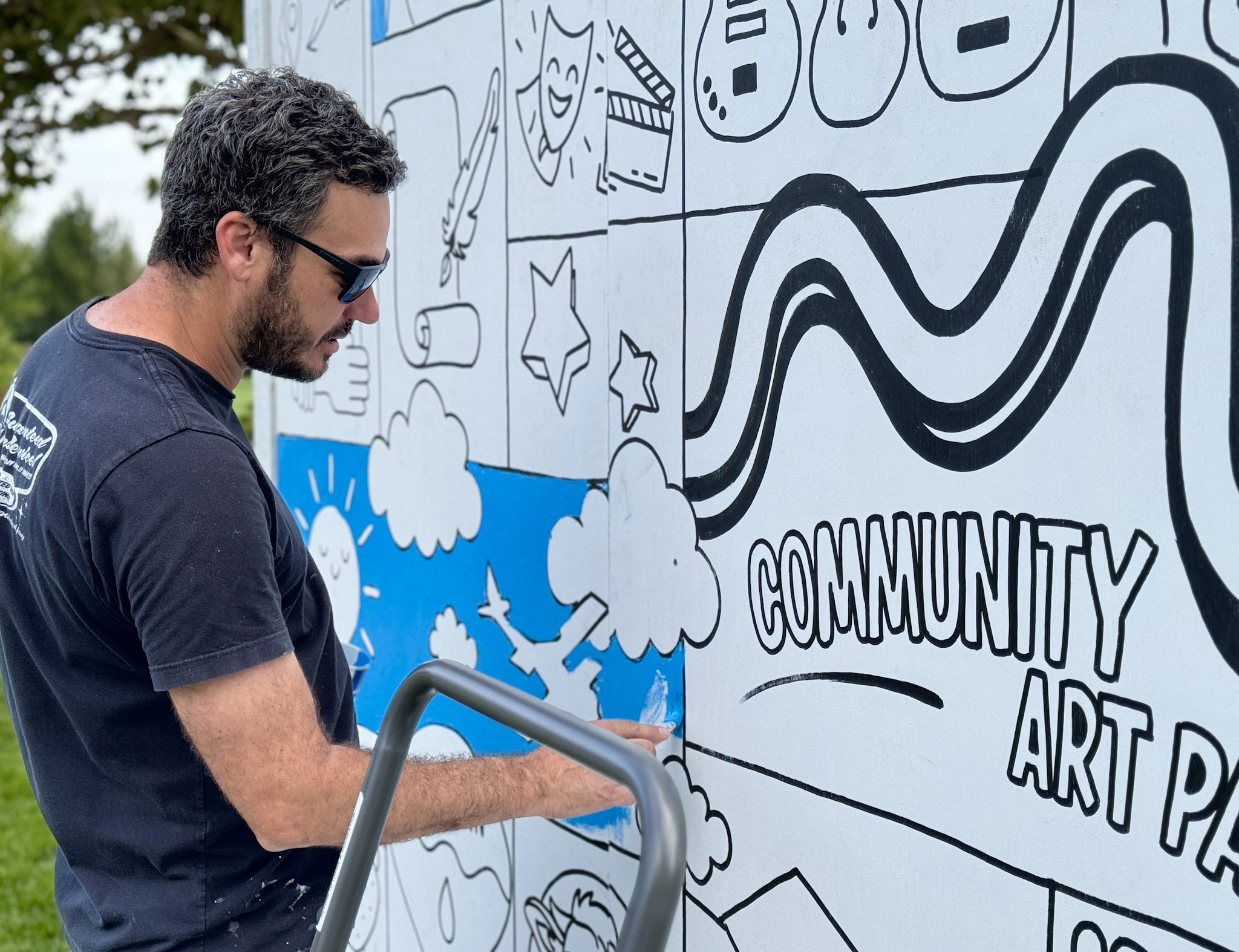Man in tshirt and sunglasses colors a blue sky in marker on a mural with the words community art party.